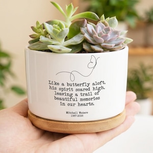 May include: A white ceramic planter with a bamboo saucer containing several succulents. The planter features a black line drawing of a butterfly and the text: Like a butterfly aloft, his spirit soared high, leaving a trail of beautiful memories in our hearts. Mitchell Waters 1987-2026.