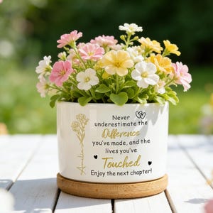 May include: A white ceramic plant pot with a wooden base, filled with colourful flowers in shades of pink, yellow, and white. The pot has text that reads "Never underestimate the Difference you've made, and the lives you've Touched. Enjoy the next chapter!"