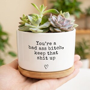 May include: A white ceramic cylindrical planter with a bamboo saucer base, containing a cluster of green and purple succulents. The front of the planter features black printed text that reads: You're a bad ass bitch, keep that shit up, with a small heart icon.