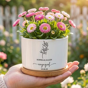 May include: A white ceramic planter with pink and white flowers, resting on a wooden saucer. The planter has the names "SOPHIA & ALLEN" and the word "engaged" with a date. The flowers are in full bloom.