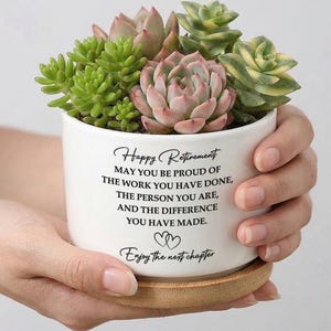 Puede incluir: Una maceta de cerámica blanca con una variedad de suculentas, incluyendo variedades verdes y rosas, se encuentra sobre un posavasos de madera. La maceta tiene el texto "Happy Retirement" y un mensaje. La maceta es sostenida por una persona.