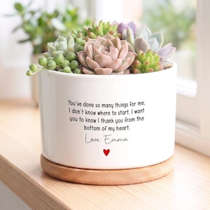 May include: A white ceramic planter containing a variety of green and pink succulents. The planter features black printed text that reads: You've done so many things for me, I don't know where to start. I want you to know I thank you from the bottom of my heart. Love. Emma, with a small red heart below.