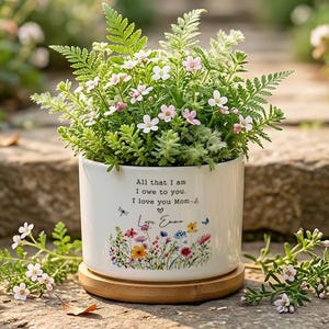 May include: A white ceramic planter filled with greenery and small pink and white flowers. The planter has a wooden base and features a floral design with the text "All that I am I owe to you. I love you Mom-".