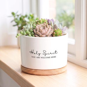 May include: A white ceramic planter filled with a variety of succulents in shades of green, pink, and purple. The planter has the words "Holy Spirit, You Are Welcome Here" in black script. It sits on a round, wooden coaster.