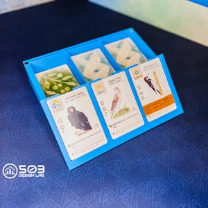 May include: A bright blue plastic card organizer tray featuring three distinct compartments. Three bird-themed game cards are displayed upright in the front slots, showing illustrations of a California Condor, a Sprague's Pipit, and an Acorn Woodpecker.