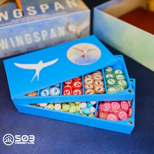 May include: A bright blue plastic board game organizer tray with a white bird silhouette and a circular bird illustration on the lid. The tray features multiple compartments filled with small round game tokens in red, green, blue, and tan, plus pastel-colored egg-shaped pieces.
