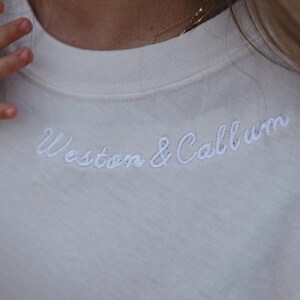 May include: Cream-coloured sweatshirt featuring the embroidered text "Weston & Callum" in white script. The image focuses on the neckline and upper chest area of the garment.