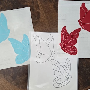 May include: Three sheets of adhesive vinyl decals featuring butterfly designs. One sheet has two light blue butterflies, one has two red butterflies, and one has two black-outlined butterfly shapes. Each butterfly contains numbered sections.