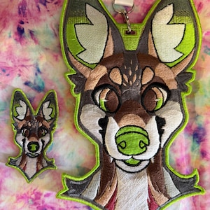 May include: Embroidered patch featuring a stylized furry character face with large eyes, a green nose, and a green outline. A smaller matching patch is also present. The patches are brown, white, and green.