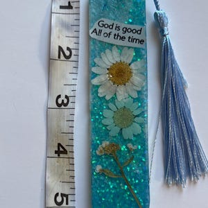 Könnte beinhalten: Handgefertigter Lesezeichen, ca. 13 cm lang, aus klarem Harz mit eingebetteten getrockneten Blumen und Glitzer. Der Lesezeichen hat eine hellblaue Quaste und den Text "God is good All of the time".