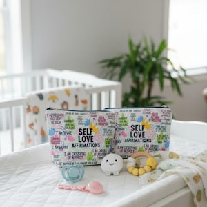 May include: Two white and colorful zippered pouches with the text "SELF LOVE AFFIRMATIONS" and other affirmations. A pacifier, a teething toy, and a plush toy are on a white changing table. A crib is in the background.