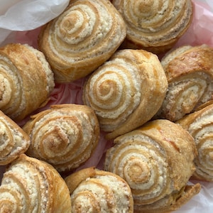 May include: A collection of freshly baked, spiral-shaped pastries. The pastries have a golden-brown exterior and a creamy, white filling, presented in a white paper bag with a pink liner. The baked goods are arranged in a close-up shot.