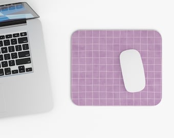 May include: A rectangular mouse pad with a light purple background and a white grid pattern. A smooth white computer mouse rests on the right side of the pad, positioned next to a silver laptop with a black keyboard.