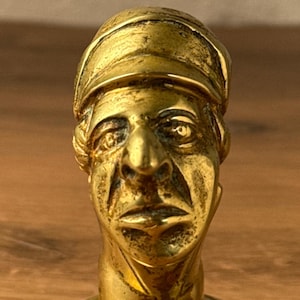 May include: A vintage brass bottle stopper featuring a sculpted bust of a man wearing a cap. The stopper has a gold finish and is set on a round base. The bust has detailed facial features.