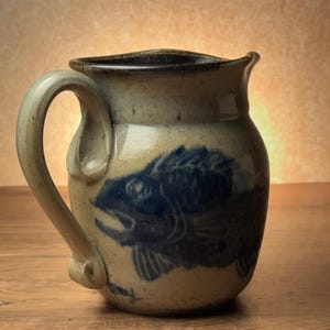 Puede incluir: Una jarra de cerámica hecha a mano con un diseño de pez azul. La jarra tiene una combinación de colores beige y marrón, con un asa curva y un pico. El pez está representado en color azul oscuro, agregando contraste al fondo claro.