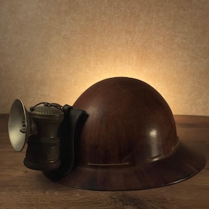 May include: A vintage brown miner's helmet with a lamp attached. The helmet is made of a dark wood-like material and has a curved brim. The lamp is metallic with a reflector and a small, detailed housing.