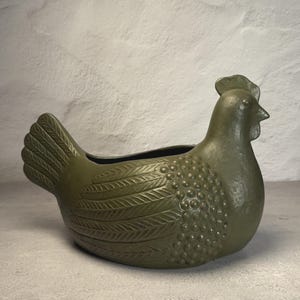 May include: An olive green ceramic planter shaped like a hen. The body features a textured surface with raised feather patterns on the wings and a series of small, rounded bumps on the chest area. The piece has a smooth, curved neck and head.