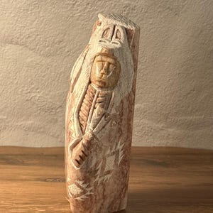 May include: A carved stone sculpture of a Native American figure. The sculpture is a light brown color with carved details of a face, headdress, and clothing. The figure is holding a staff or weapon. The sculpture is standing upright.