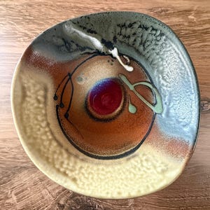 以下が含まれることがあります： 抽象的なデザインの手作りセラミックプレート。中央の赤い円は、茶色、タン、青の色合いで囲まれ、黒と緑のアクセントがあります。リムはテクスチャード加工され、斑点のある外観です。