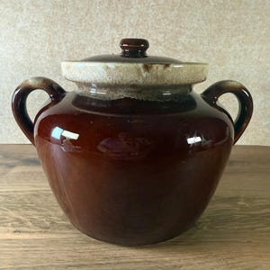 May include: A brown ceramic crock with a lid and two handles. The crock has a glossy finish and a light tan band around the top. The lid has a small, round handle. The crock is sitting on a wooden surface.