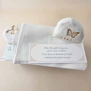 May include: Two off-white, smooth, rounded stones with butterfly impressions, one featuring the word "rest" in lowercase letters. A white fabric drawstring pouch holds a card with the text "May this gift bring you peace and comfort."