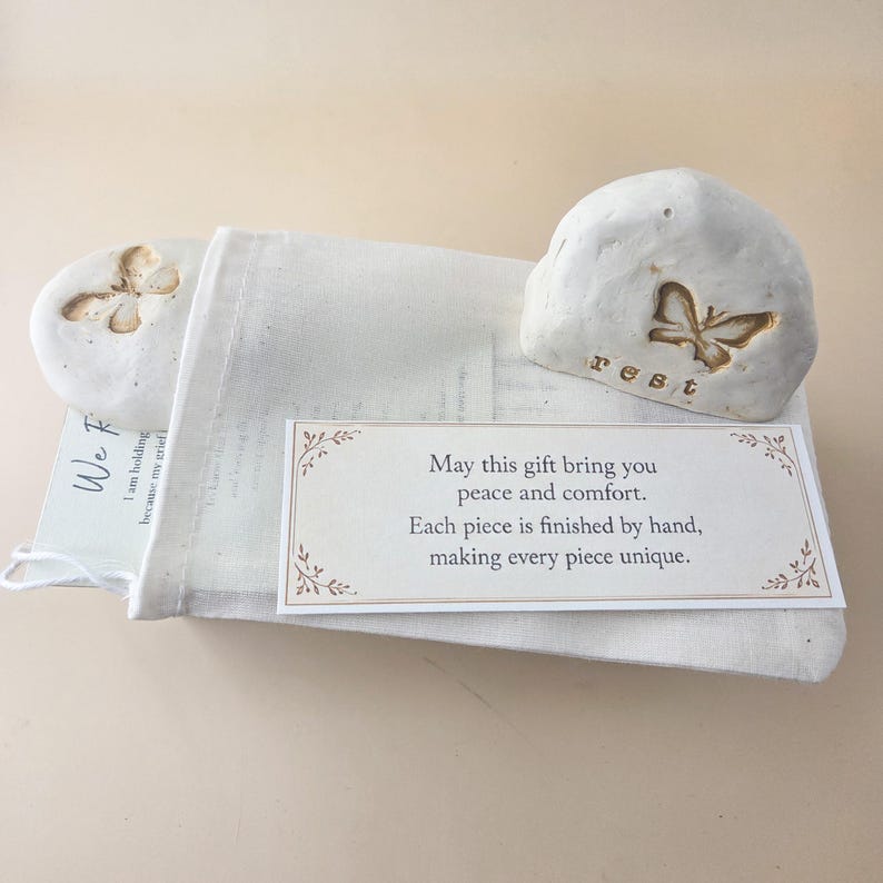 May include: Two off-white, smooth, rounded stones with butterfly impressions, one featuring the word "rest" in lowercase letters. A white fabric drawstring pouch holds a card with the text "May this gift bring you peace and comfort."