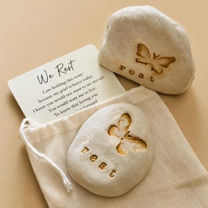 May include: Two off-white stones with engraved butterfly designs and the word rest, accompanied by a white card featuring the text We Rest and a poem about grief, all resting on a light beige fabric drawstring pouch with a white cord.