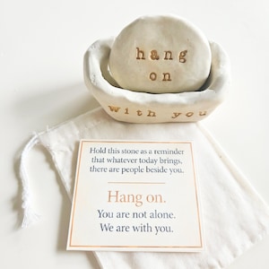 May include: A white ceramic bowl containing a smooth white stone with the text hang on stamped in brown. The bowl features the text with you stamped on the front. A white fabric drawstring bag sits beneath a square card with printed text.