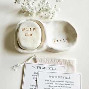 May include: A cream-colored ceramic dish with two sections, one holding a stone inscribed with "with me" and the other with "still" stamped inside. Below are two white cards with the text "WITH ME STILL" and a small drawstring pouch.