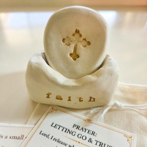May include: A small, off-white, hand-formed clay object featuring a circular disc with an embossed cross design resting in a base. The base has the word faith stamped in gold lettering. A small card with the text PRAYER: LETTING GO & TRUST is visible.
