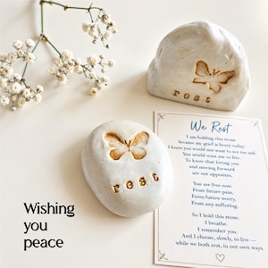 May include: Two off-white stones with embossed butterfly designs and the word rest, accompanied by a card featuring the text We Rest and a poem about grief, alongside a sprig of white baby's breath flowers and the text Wishing you peace.