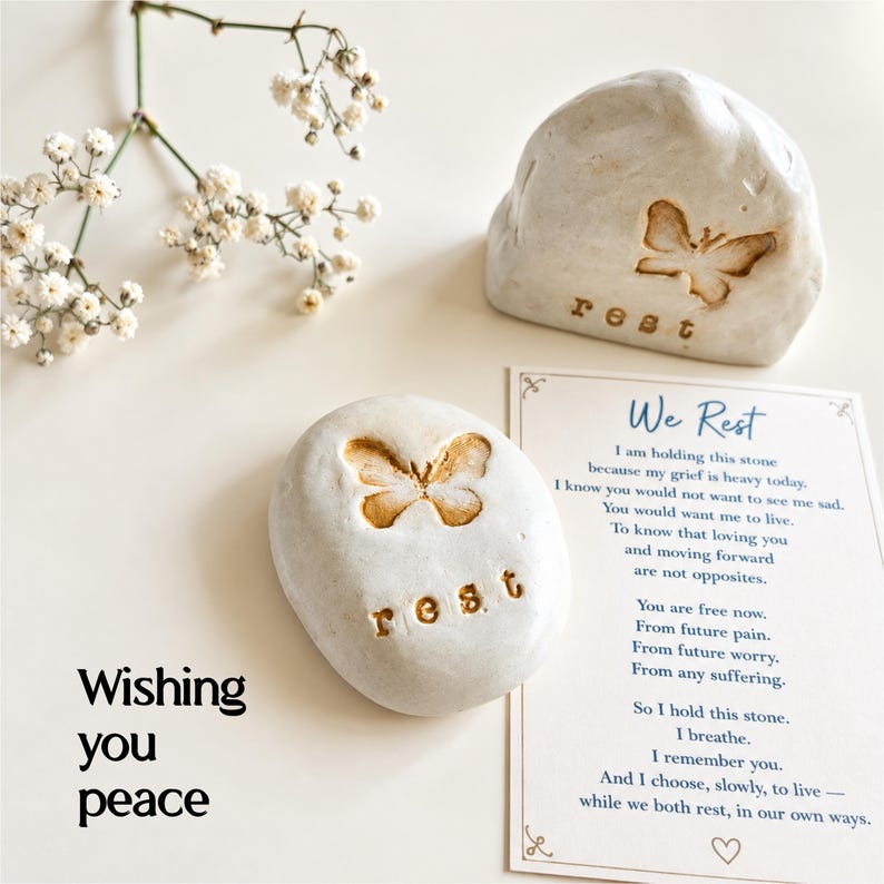 May include: Two off-white stones with embossed butterfly designs and the word rest, accompanied by a card featuring the text We Rest and a poem about grief, alongside a sprig of white baby's breath flowers and the text Wishing you peace.