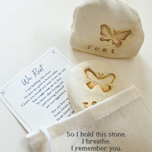May include: Two off-white stones with embossed butterfly designs and the word rest, accompanied by a white card with the text We Rest and a poem, plus a small beige fabric drawstring pouch with the text So I hold this stone. I breathe. I remember you.