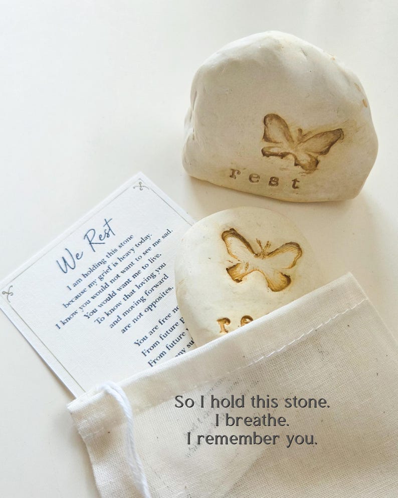 May include: Two off-white stones with embossed butterfly designs and the word rest, accompanied by a white card with the text We Rest and a poem, plus a small beige fabric drawstring pouch with the text So I hold this stone. I breathe. I remember you.