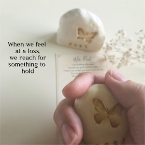 May include: A smooth, light-colored stone featuring an engraved butterfly and the text "rest" at the bottom. The stone is held in a hand, with a matching stone and a printed card with the text "We Rest" visible in the background.