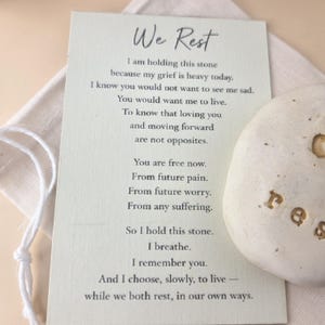 May include: A light green rectangular card with the black printed text We Rest and a poem about grief, accompanied by a smooth, off-white stone with the word rest engraved in gold, all resting on a small, light-colored fabric drawstring pouch.