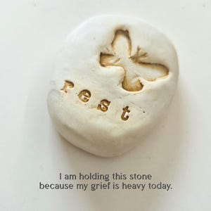 May include: An off-white, smooth, rounded stone featuring an embossed butterfly and the word "rest" in lowercase letters. Below the stone, the text reads: "I am holding this stone because my grief is heavy today." The object has a matte finish.