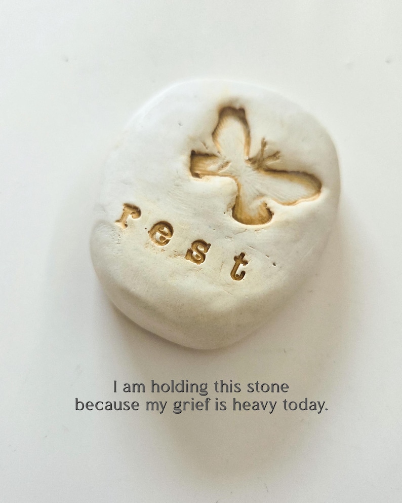 May include: An off-white, smooth, rounded stone featuring an embossed butterfly and the word "rest" in lowercase letters. Below the stone, the text reads: "I am holding this stone because my grief is heavy today." The object has a matte finish.