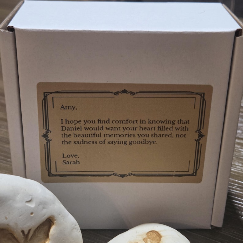 May include: A white cardboard box featuring a rectangular tan label with a black decorative border. The text reads: Amy, I hope you find comfort in knowing that Daniel would want your heart filled with the beautiful memories you shared, not the sadness of saying goodbye. Love, Sarah.