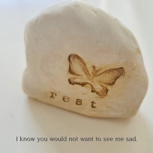 May include: A smooth, off-white stone with an irregular, rounded shape. The surface features an embossed butterfly silhouette and the lowercase text "rest" stamped below it. The text "I know you would not want to see me sad." appears below the stone.