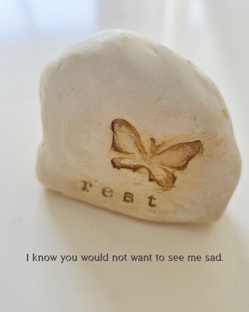 May include: A smooth, off-white stone with an irregular, rounded shape. The surface features an embossed butterfly silhouette and the lowercase text "rest" stamped below it. The text "I know you would not want to see me sad." appears below the stone.