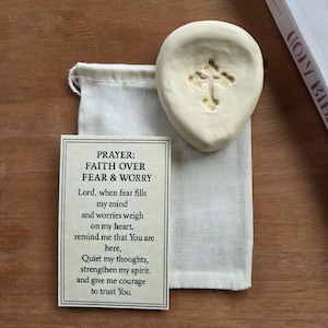 May include: A cream-colored, cross-shaped stone sits atop a small, off-white drawstring bag. A card with the text "PRAYER: FAITH OVER FEAR & WORRY" is visible. The prayer text is printed in black. The background is a wooden surface.