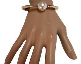 May include: Gold-tone bracelet featuring a large, round, white pearl. The bracelet has a simple, elegant design with a thick band. The pearl is centered on the bracelet, creating a focal point. This jewelry piece is suitable for various occasions.