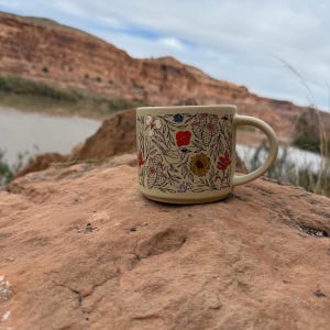 May include: A beige ceramic mug with a floral pattern featuring red, yellow, and blue flowers, and green cacti. The mug is set on a reddish-brown rock with a scenic background of a river and mountains.
