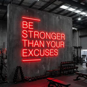 Op de afbeelding: Een rood neonbord op een grijze betonnen muur leest "BE STRONGER THAN YOUR EXCUSES." Het bord staat in een sportschool, met gewichten en trainingsapparatuur zichtbaar op de achtergrond. De vloer is rood.