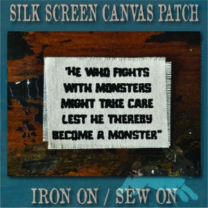 May include: A rectangular off-white canvas patch with frayed edges featuring black screen-printed text that reads: HE WHO FIGHTS WITH MONSTERS MIGHT TAKE CARE LEST HE THEREBY BECOME A MONSTER. The patch is designed for iron-on or sew-on application.
