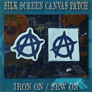 May include: Two off-white canvas patches featuring a black silk-screened anarchy symbol. One patch is cut to the shape of the symbol, while the other is a square with frayed edges. The text SILK SCREEN CANVAS PATCH and IRON ON / SEW ON is visible.