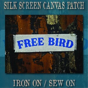 May include: A rectangular light grey canvas patch with frayed edges featuring the text FREE BIRD printed in bold blue ink. The patch is displayed against a dark brown and black textured background with gold accents.