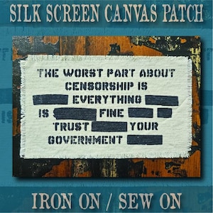 May include: A rectangular off-white canvas patch with frayed edges featuring black screen-printed text that reads: THE WORST PART ABOUT CENSORSHIP IS EVERYTHING IS FINE TRUST YOUR GOVERNMENT, with several words obscured by solid black bars.