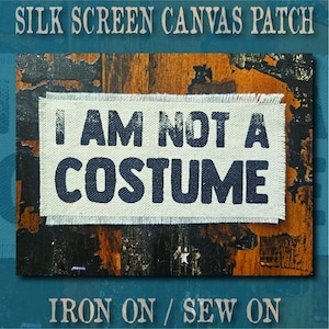 May include: A rectangular off-white canvas patch with frayed edges featuring the black screen-printed text I AM NOT A COSTUME. The patch is displayed against a dark, weathered wood background with visible orange and black paint textures.
